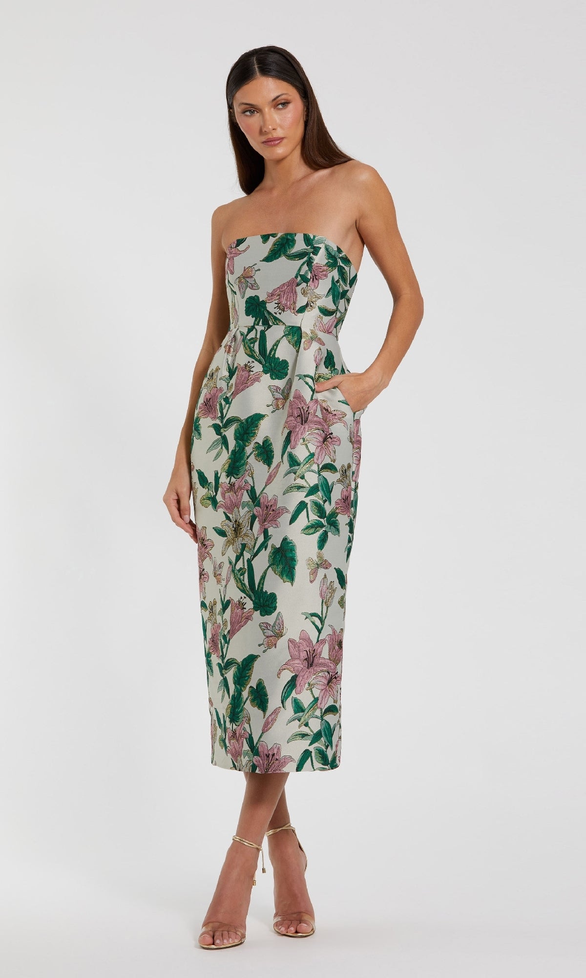 A woman stands wearing the Mac Duggal 56097 Midi Floral Wedding Guest Dress, a strapless midi dress with green and pink floral accents, paired with light high-heeled sandals against a plain white background.